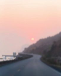 This is a soft, dreamy depiction of a sunset sky over a winding coastal highway, rendered in an impressionistic style. The sc
