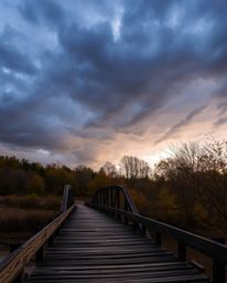 This photograph captures an old wooden bridge over a river at sunset during a period of intense autumn weather. The scene is 