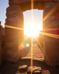 A bright lens flare illuminates a scene where a sunset casts shadows through ancient stone ruins. The image features natural 