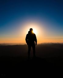 Witness a powerful scene from a high alpine peak, where a silhouetted figure stands against the sky. The bright sunset provid