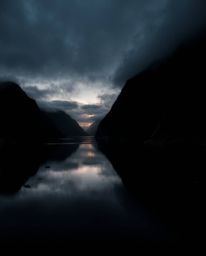 A moody high-angle photograph depicting a glassy fjord reflecting a sunset. Captured from an elevated, looking-down perspecti