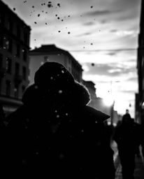 An intense black and white candid street photograph captures the dynamic energy of snowflakes falling during a vibrant sunset