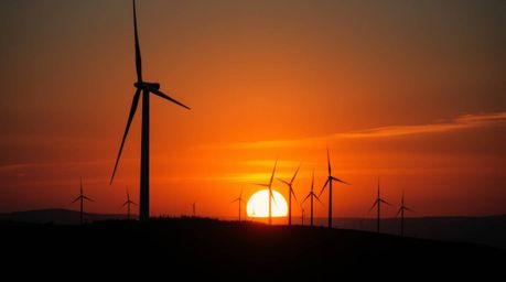 A high-resolution stock photo depicting a dramatic sunset behind a row of wind turbines on a hill. The image utilizes dark to