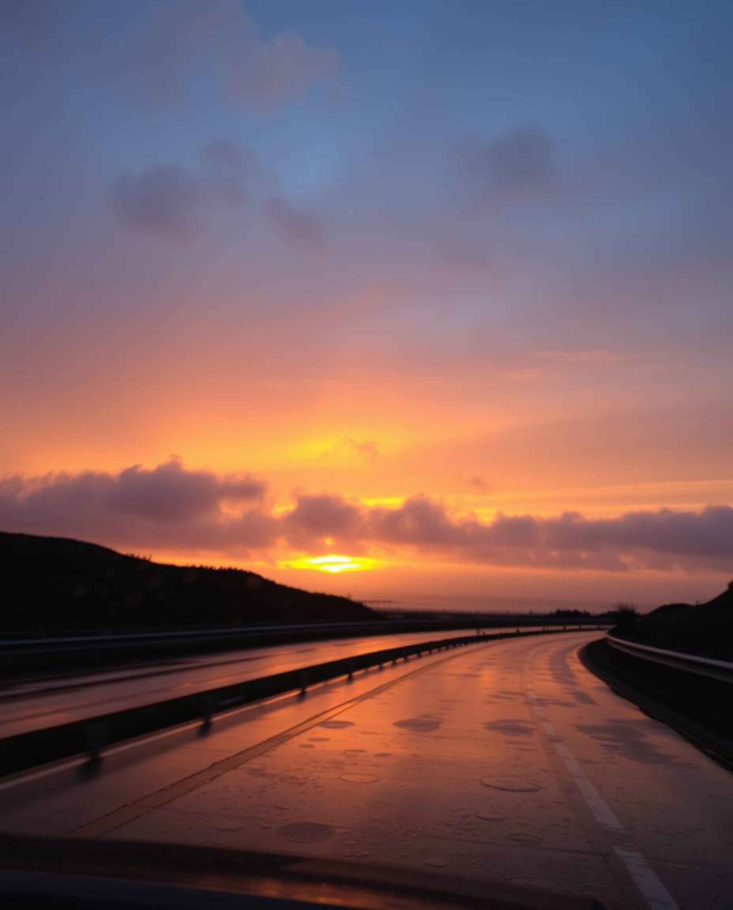 This image captures a beautifully melancholic scene of a sunset sky over a winding coastal highway under wet, rainy condition