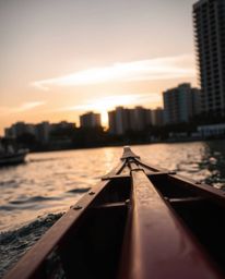 A candid close-up street photograph captures a boat drifting on a lake at sunset, with a visible urban background creating a 