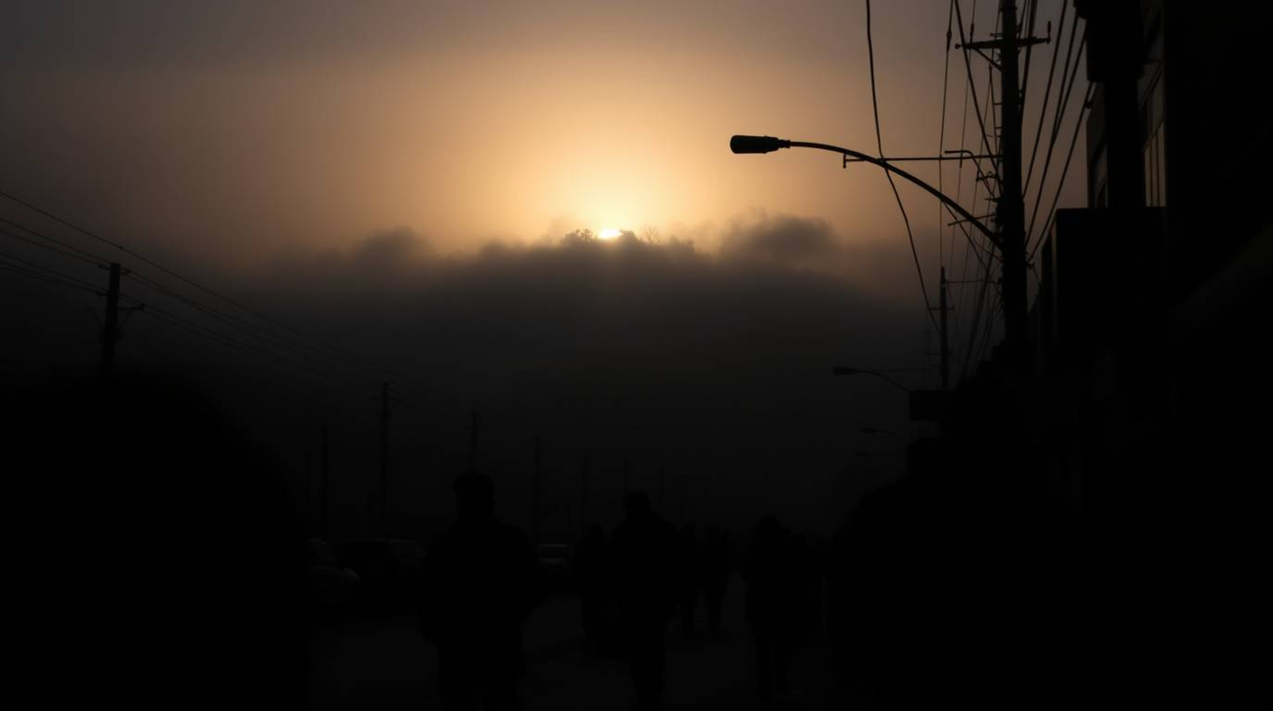 An intense, jet-black candid street photograph capturing the atmospheric beauty of fog-covered hills as the sunset attempts t