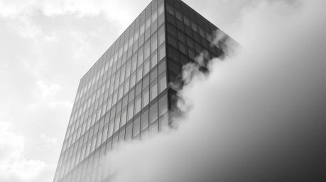 A captivating black and white photograph showcases a modern glass building seemingly dissolving into the soft clouds above, c
