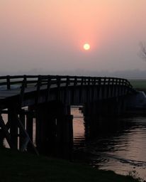 A precise architectural color photograph captures an old wooden bridge over a river at sunset. The image is taken from a wide