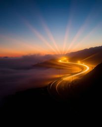 Stunning volumetric lighting illuminates a winding coastal highway at sunset, creating a scene of breathtaking beauty. Visibl