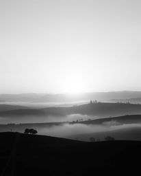 This is a timeless and elegant black and white photograph depicting a sunset over the misty, rolling hills of Tuscany. The im
