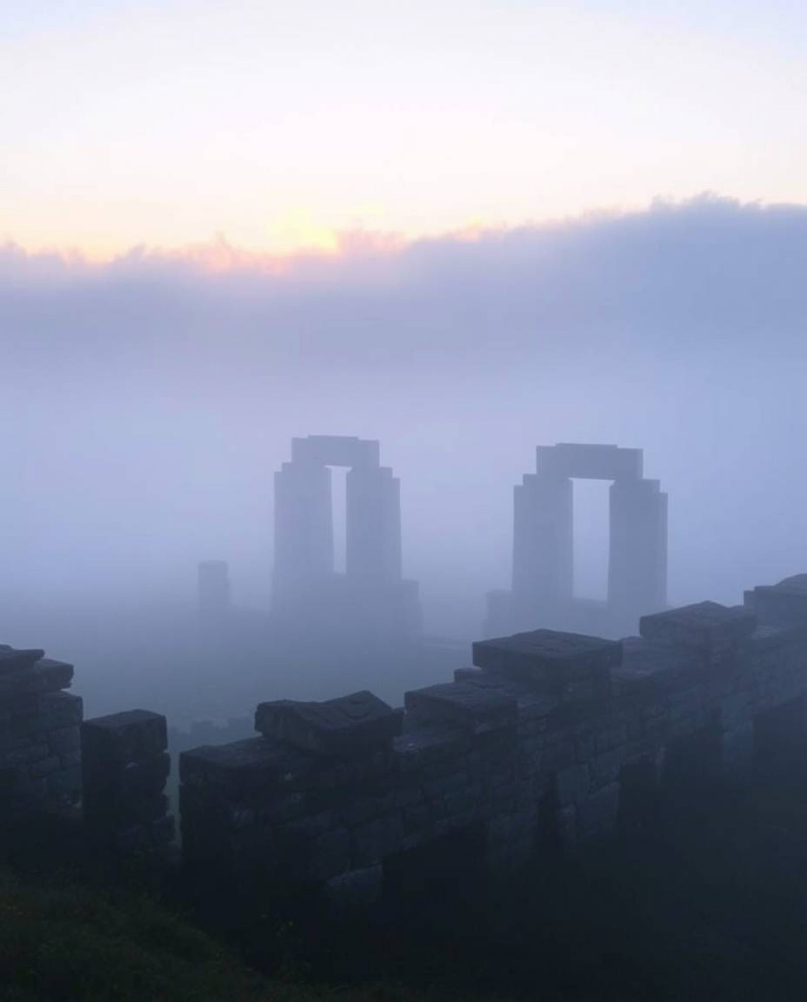 Thick fog surrounds ancient stone ruins at sunset, casting deep shadows and creating a mysterious atmosphere. The scene is en