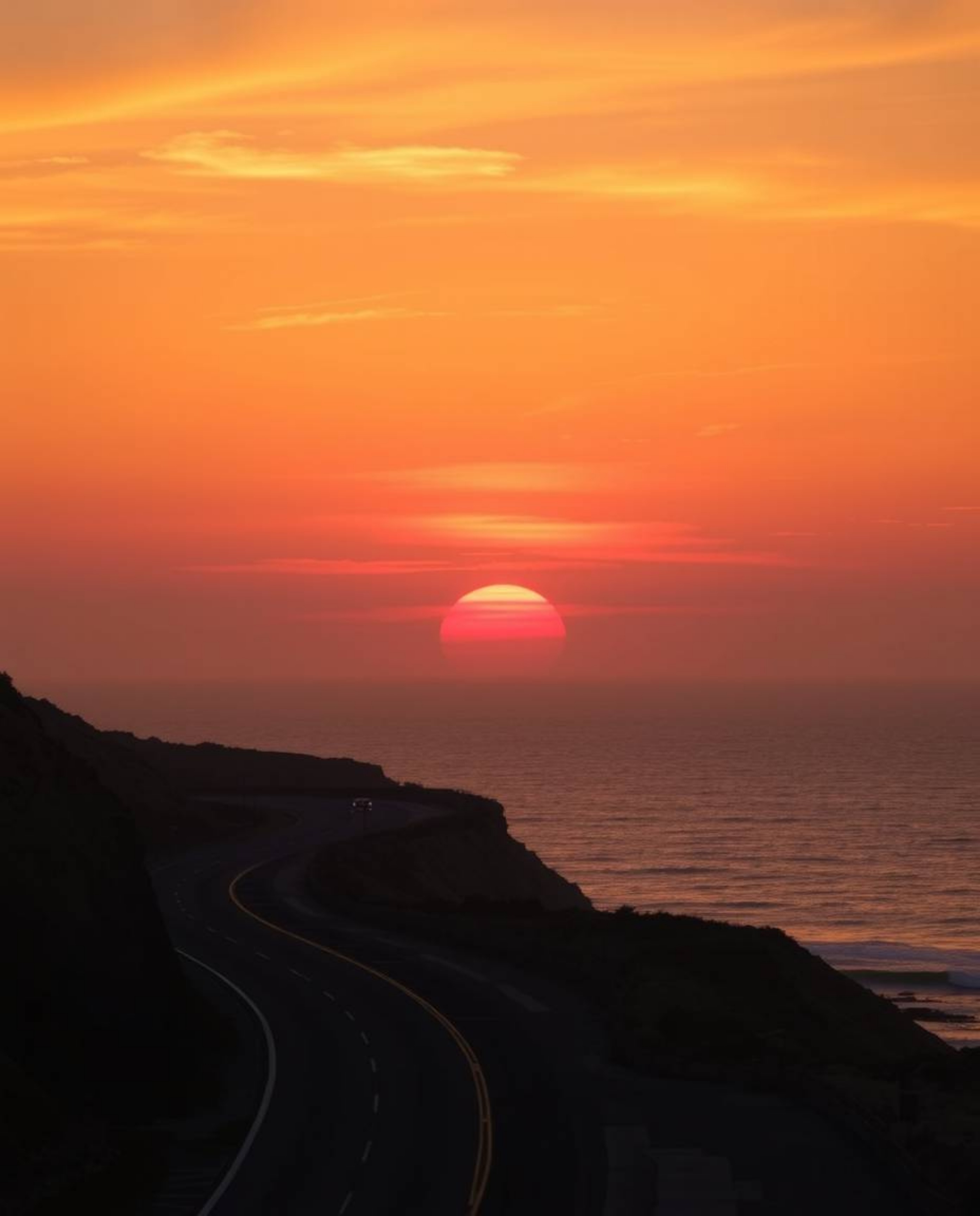 This is a professional fine art photograph of a warm sunset sky over a winding coastal highway. The scene is dominated by a p
