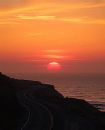 This is a professional fine art photograph of a warm sunset sky over a winding coastal highway. The scene is dominated by a p