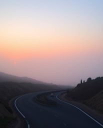 This wide shot captures a soft and dreamy sunset sky over a winding coastal highway. The scene is enveloped in hazy, soft lig