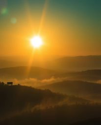 Photograph of a sunset over misty Tuscan hills. Rich deep teal, bright lens flare, light streaks, vibrant colors. Luxurious, 
