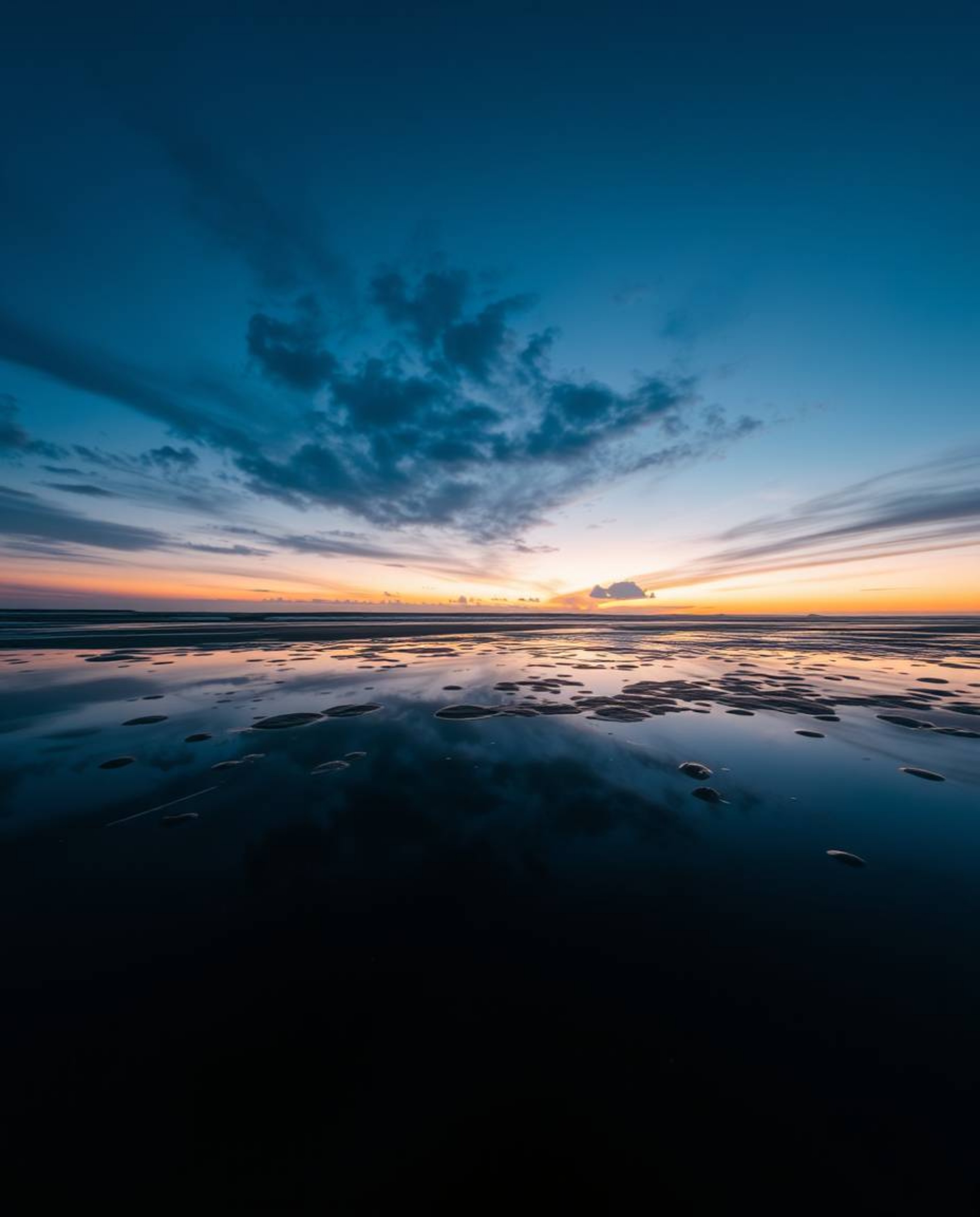 A high contrast, low key photograph captures a sunset sky as it is mirrored in the tidal pools on a beach. The image is defin