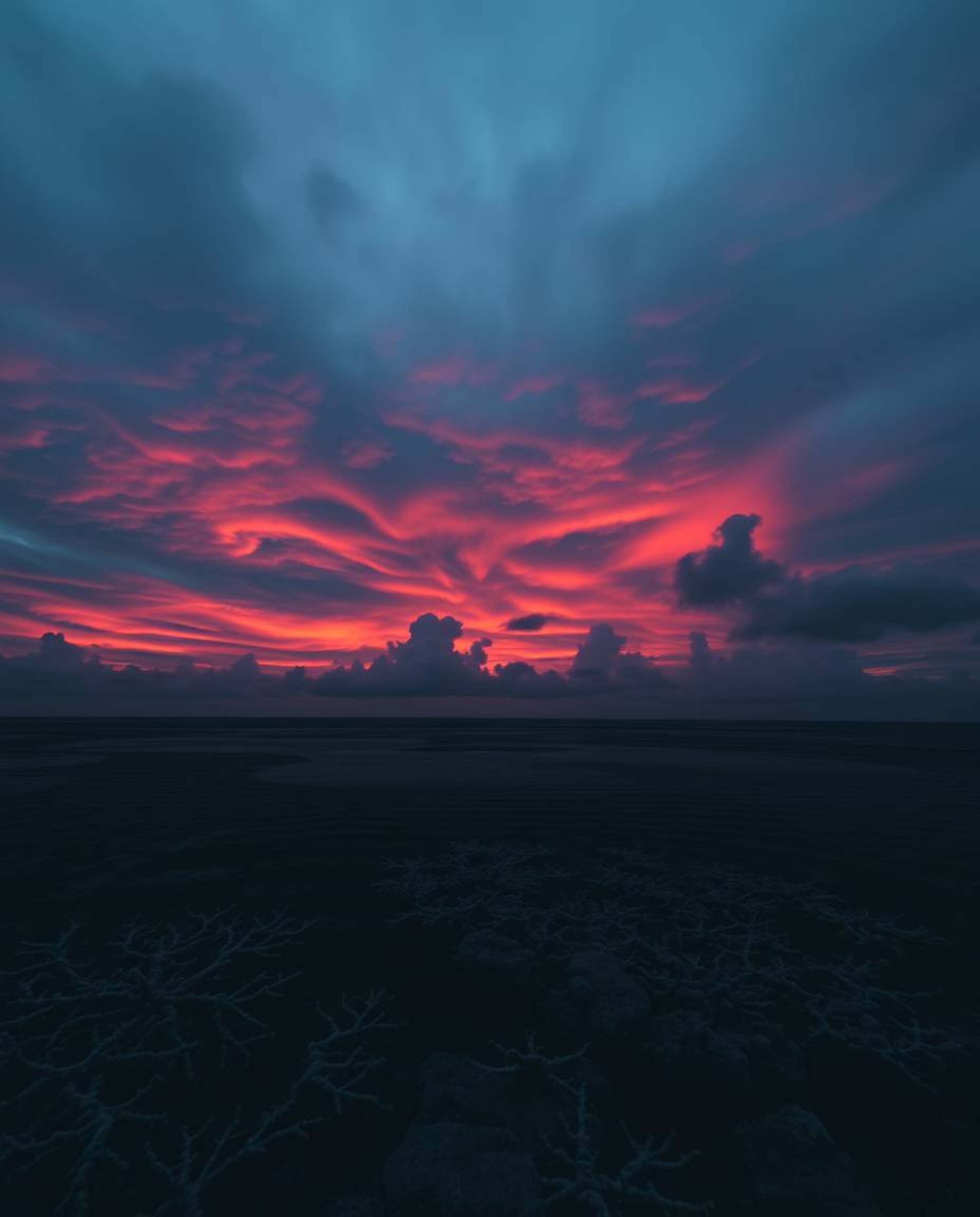 A moody image of a colorful sky over a coral reef at low tide, rendered in dark tones and low contrast. This piece is charact