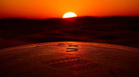 A warm cast bronze surface is silhouetted against the backdrop of a sunset over golden desert dunes. The piece boasts an anti