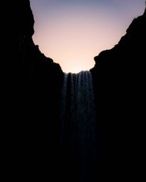 A low-angle, high-contrast photograph with split lighting showcases a dramatic sunset behind a waterfall. This powerful compo