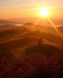 Photograph of a sunset over misty Tuscan hills. Warm autumn colors, bright lens flare, light streaks, vibrant colors. Earthy,