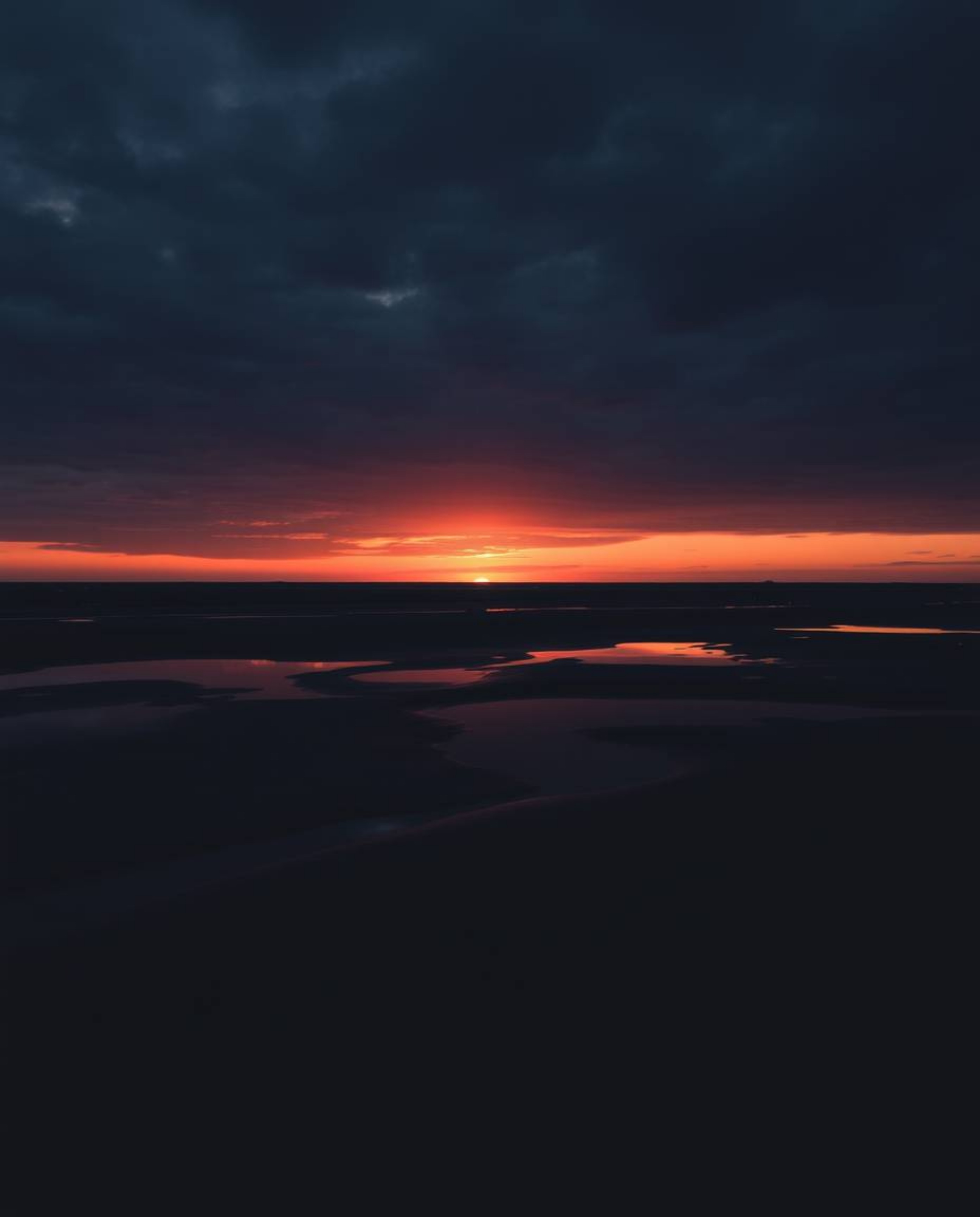 This is a moody, low-contrast image of a sunset sky mirrored in tidal pools on a beach. The artwork features a strong style t