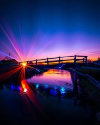 An old wooden bridge over a river at sunset is reimagined in striking, deep ultraviolet hues. The scene is characterized by v