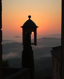 This is an architectural color photograph depicting a sunset over the misty, rolling hills of Tuscany. The image is character