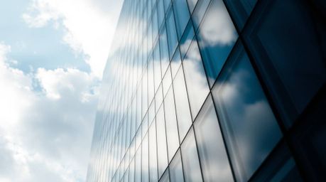 A close-up shot bathed in soft, gentle light captures a modern glass building seemingly dissolving into a sky where soft clou