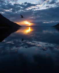 A professional fine art photograph of a glassy fjord reflecting a sunset. This image masterfully employs chiaroscuro lighting