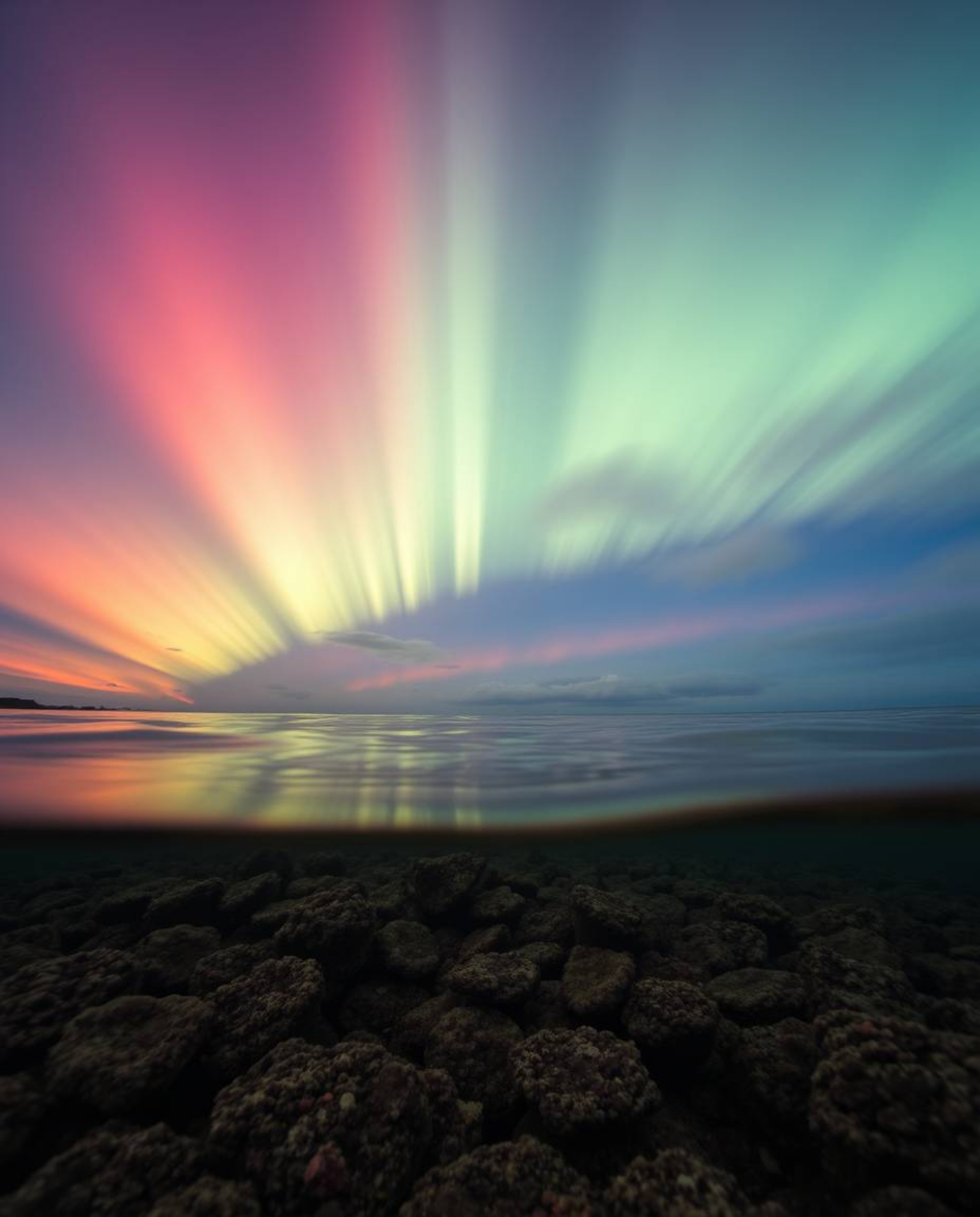 A stunning long exposure photograph captures a colorful sky over a coral reef at low tide from a dynamic low-angle perspectiv