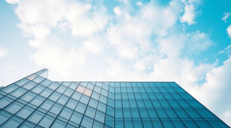 A precise architectural color photograph capturing a modern glass building seemingly dissolving into a soft, cloud-filled sky