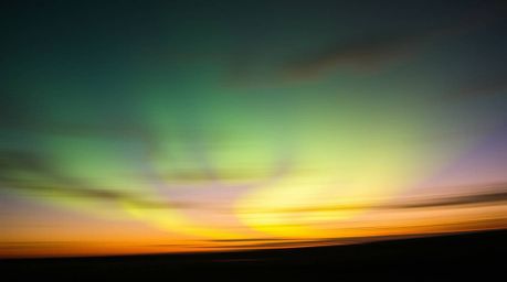 A captivating editorial-style photograph showcases the dynamic emergence of the Northern Lights over a vibrant sunset sky.  T