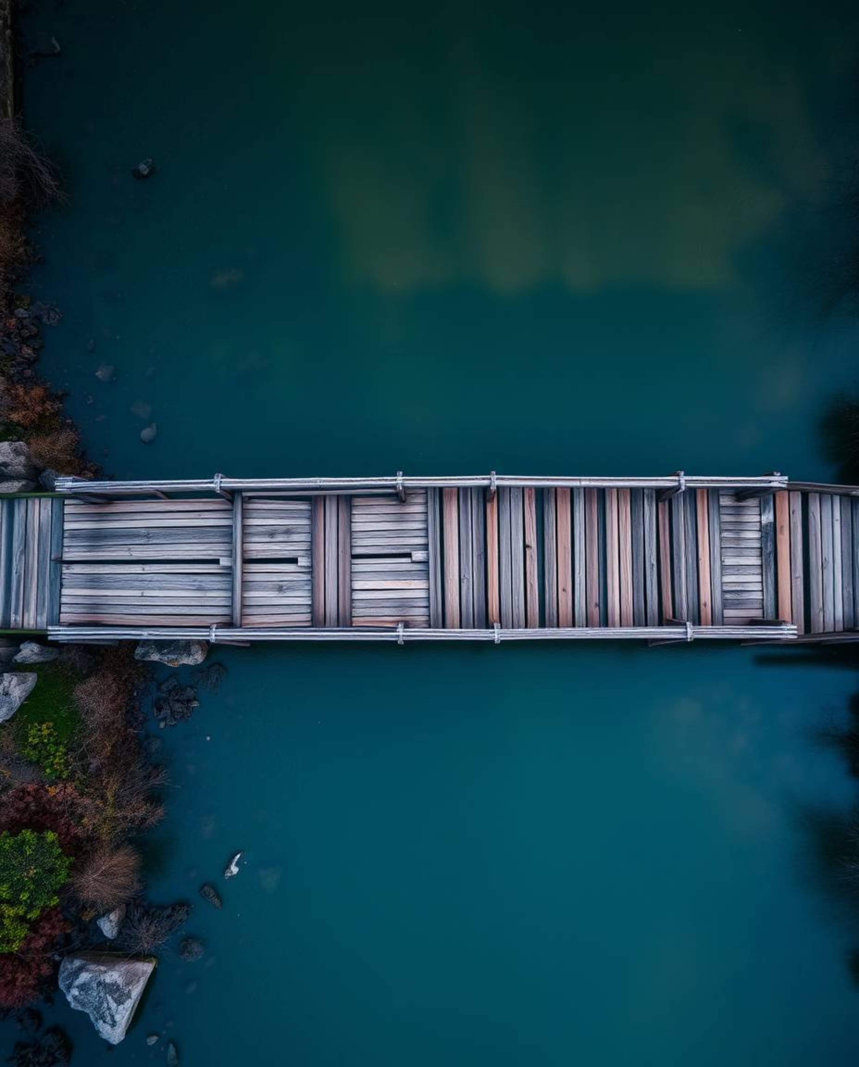 An overhead bird's-eye view of an old wooden bridge over a river at sunset. Captured from a top-down perspective, the image i
