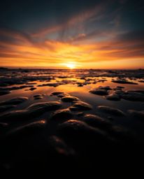 A tight close-up shot, defined by contrasting chiaroscuro lighting, depicts a sunset sky mirrored in the still waters of tida