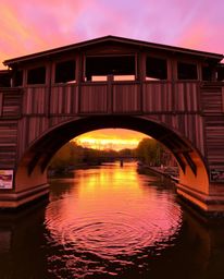 A photograph depicts a warm-toned old wooden bridge arching gracefully over a river at sunset. The scene is filled with the r