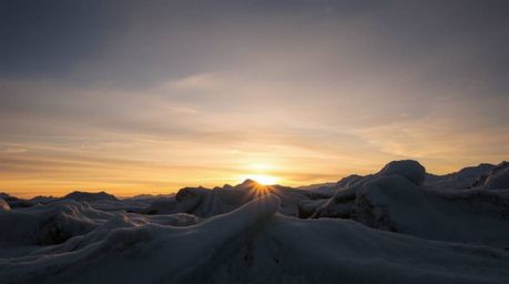 A breathtaking low-angle photograph captures a dramatic sunset casting warm earth tones, browns, and beiges across icy glacie