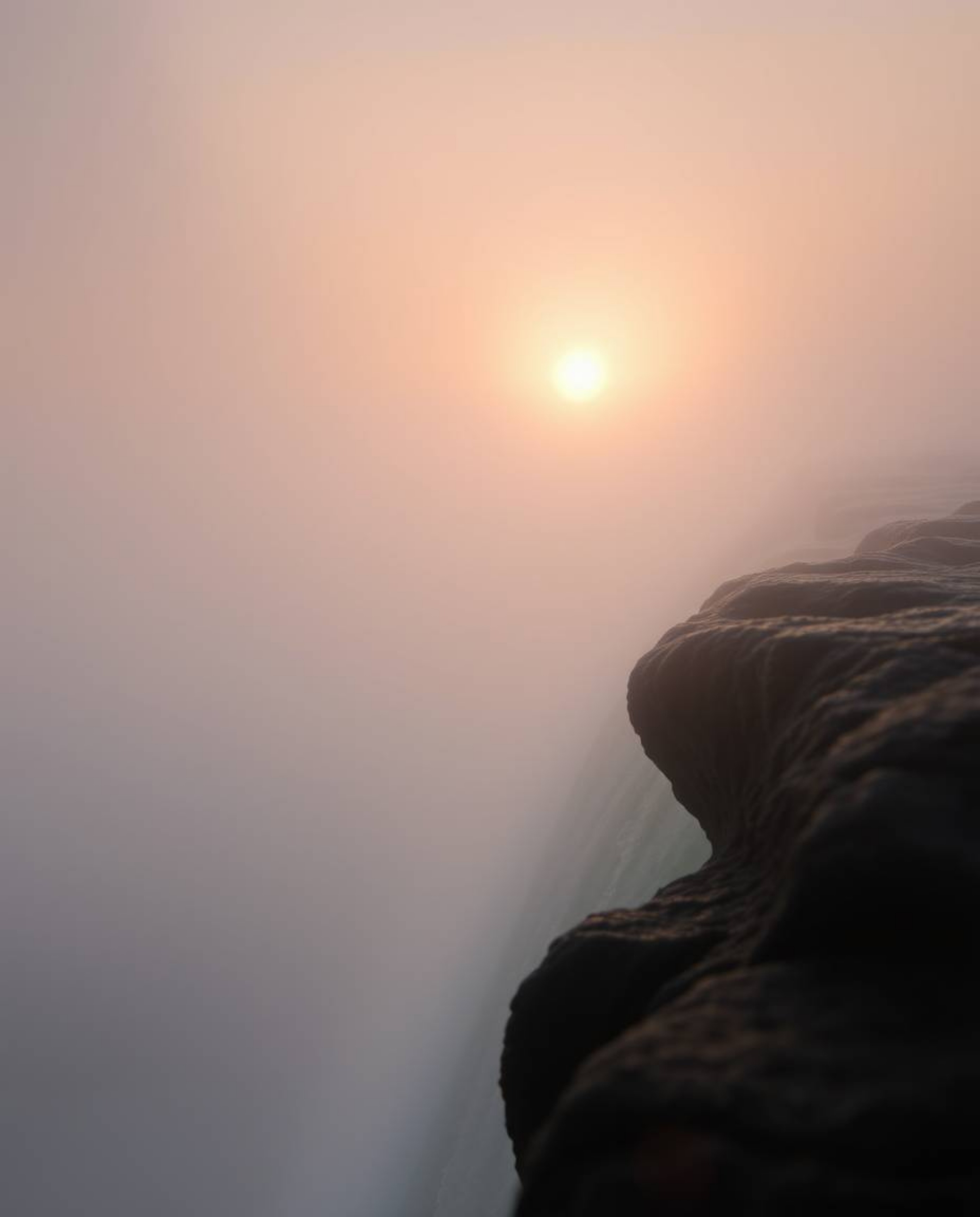 An extreme macro close-up captures a sun setting behind a dramatic waterfall amidst thick foggy surroundings. The scene is ch