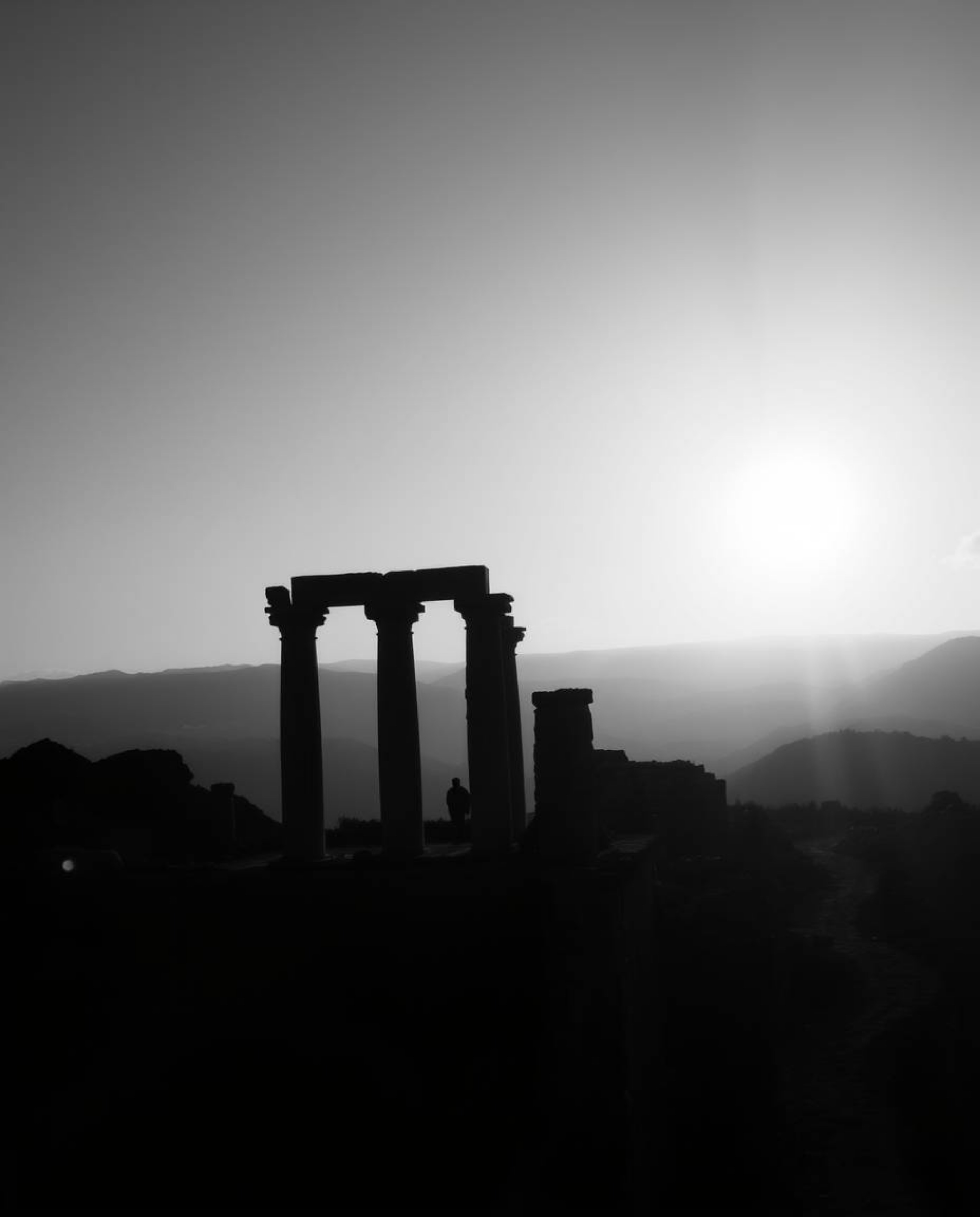 An overhead shot, presented as a classic black and white photograph, captures ancient stone ruins silhouetted against a sunse