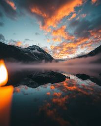 A dramatic, low-angle shot captures the stunning reflections of fiery clouds in a mountain lake. The scene is softly illumina