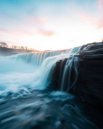 A dynamic long-exposure photograph of a waterfall at sunset, emphasizing motion blur and light trails. The image captures act