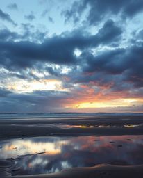 A graceful and elegant sunset sky is mirrored in the tidal pools of a beach, set against a backdrop of intense dramatic weath