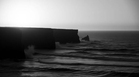 A classic black and white photograph captures the timeless elegance of waves crashing against cliffs under a dramatic sunset 
