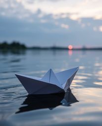 This is a tight close-up shot that captures an intricately folded paper boat drifting on a serene lake at sunset. The artwork