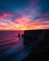 A stunning long-exposure photograph captures the motion and light trails of a sunset over a coastal cliff. The soaring birds 