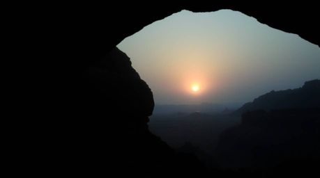 Experience the serene beauty of a desert sunset framed by a natural rock arch in this moody, low-contrast image. The scene is