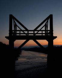 An intense jet black logo of an old wooden bridge over a river at sunset. This design is composed of geometric shapes and cle