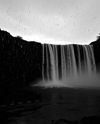 A classic black and white photograph of a rainy scene, featuring a waterfall at sunset. Drizzling rain creates reflective sur