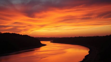 This stunning fine art photograph captures a winding river bathed in the warm glow of a dramatic orange-pink sunset.  Profess