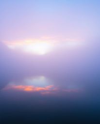 A mysterious landscape shrouded in thick fog, dominated by a cool color palette of blues, greens, and purples. The low visibi