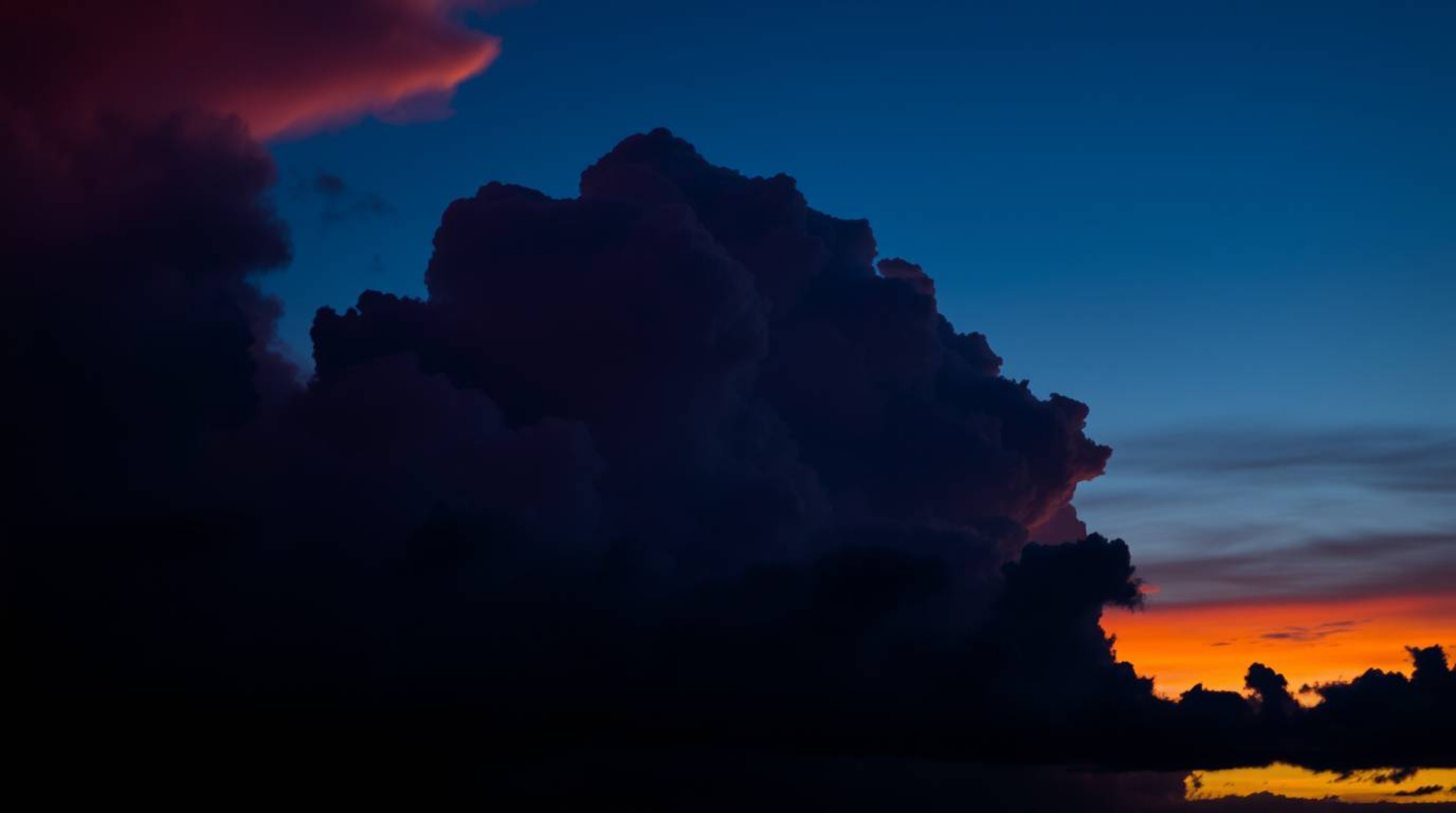 Dramatic silhouette of thunderstorm clouds, backlit by a vibrant sunset.  The image features rich burgundy and deep blue tone